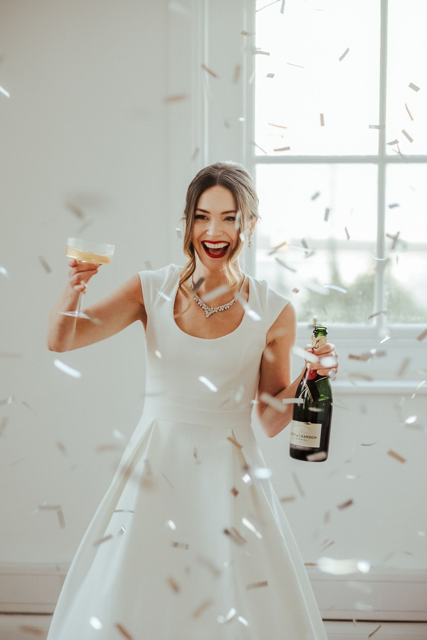 Person in white dress celebrates joyfully with champagne as confetti falls in bright interior space.