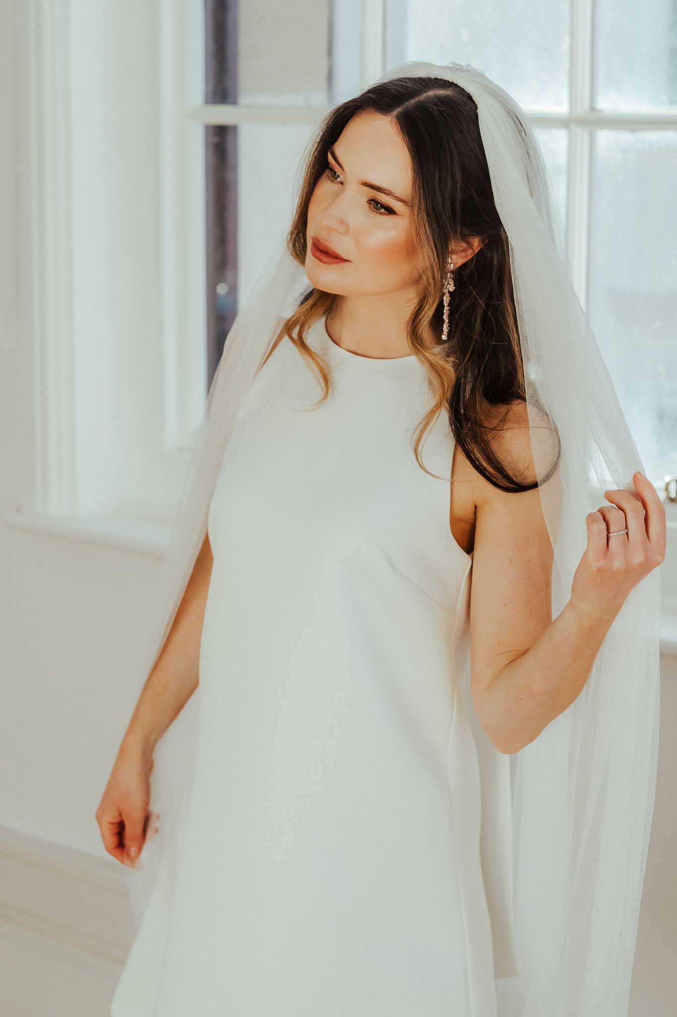 A bride in a minimalist white wedding dress and veil poses elegantly by a window in a bright interior setting.