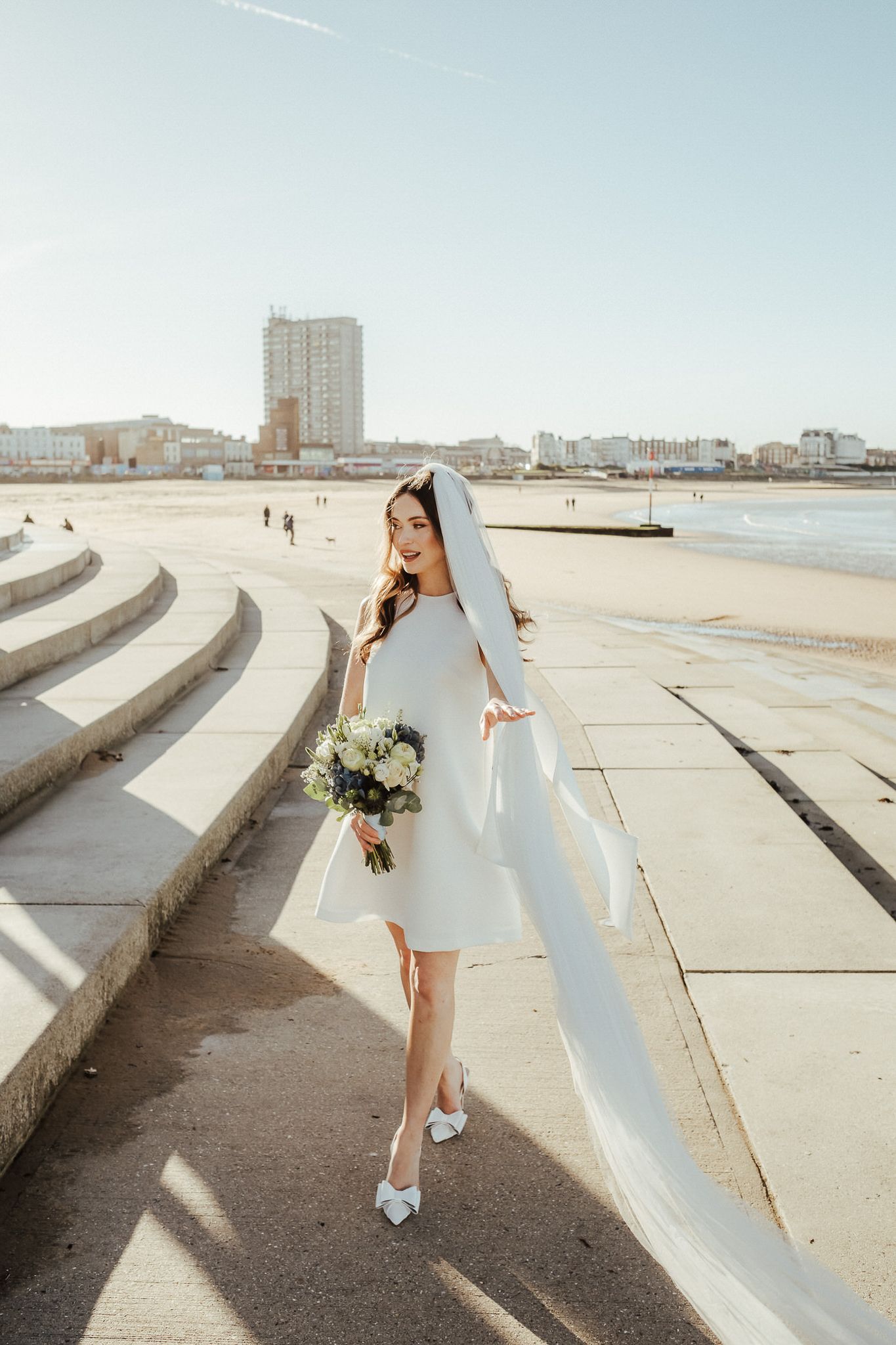 A person in a short white dress poses with a bouquet on concrete steps along a beachfront.