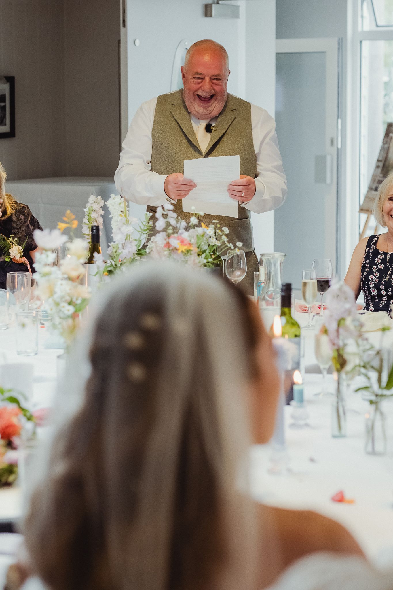 A speaker delivers a heartfelt speech during an elegant wedding reception with white floral centerpieces and wine glasses.
