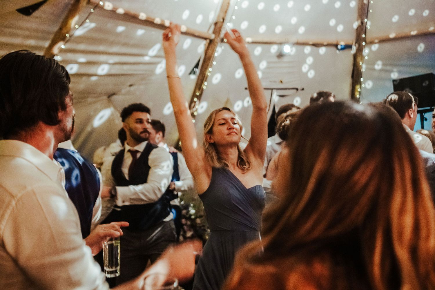 Wedding guests dancing and celebrating under string lights in a tent venue.