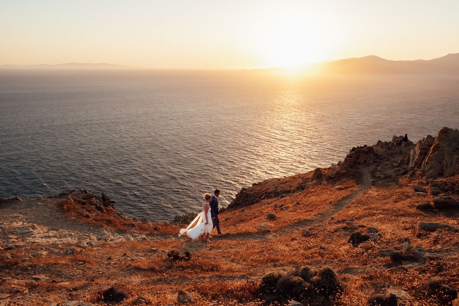 Bride and groom standing on rocky coastal cliffside at golden sunset with ocean and mountain views.