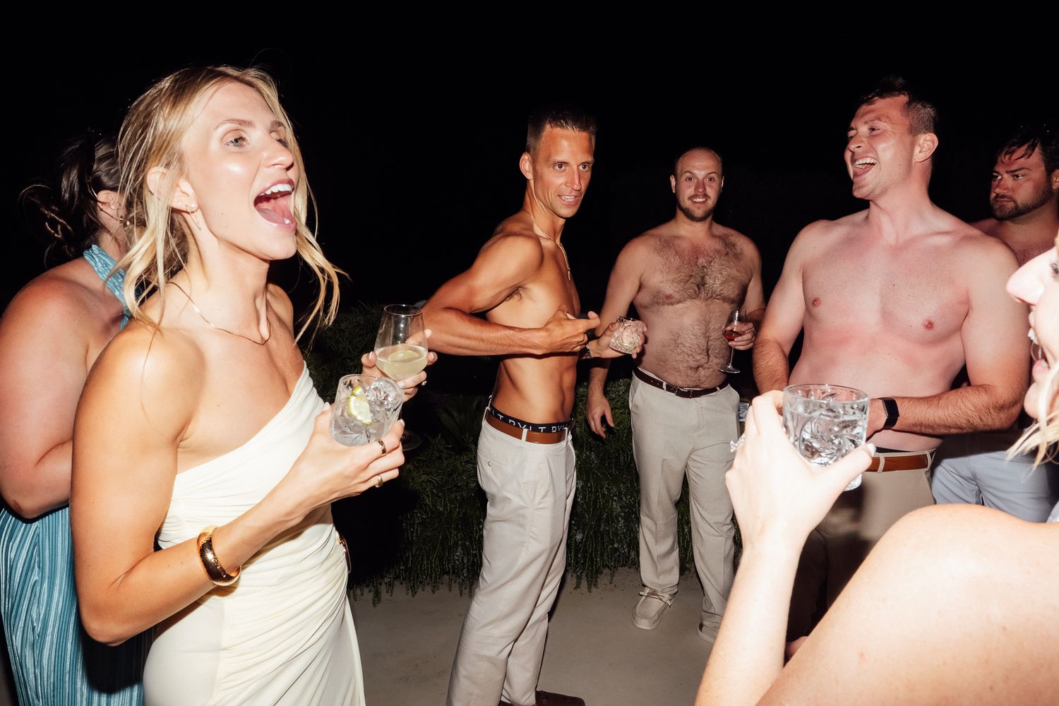 Group of friends laughing and celebrating at an outdoor nighttime party, some shirtless men holding drinks.