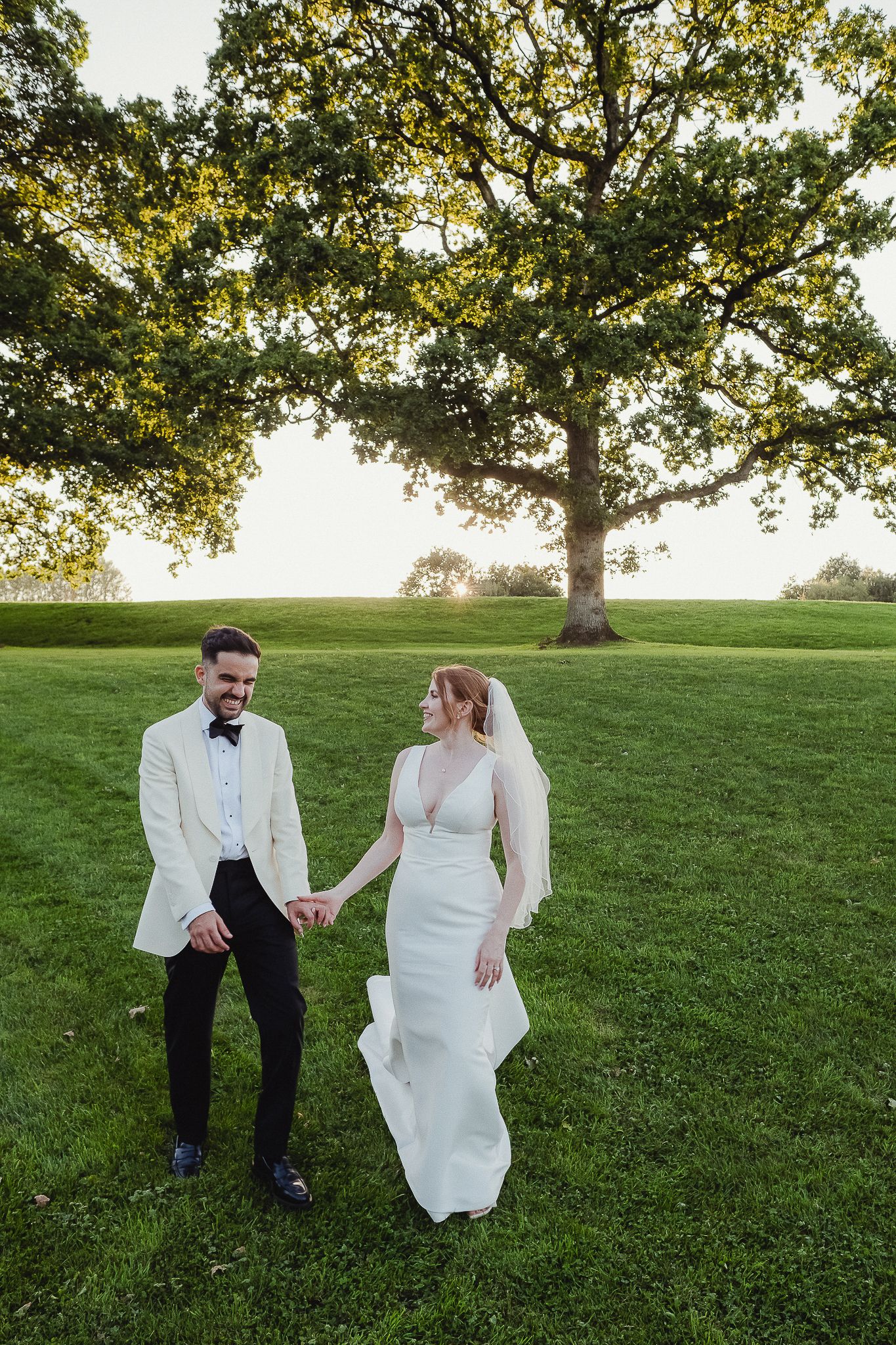 A couple in wedding attire shares a playful moment while sitting on a grassy hill under a large tree.