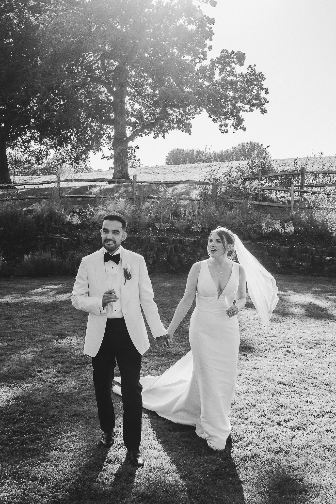 A couple in wedding attire runs playfully through a sunlit garden setting in black and white photography.