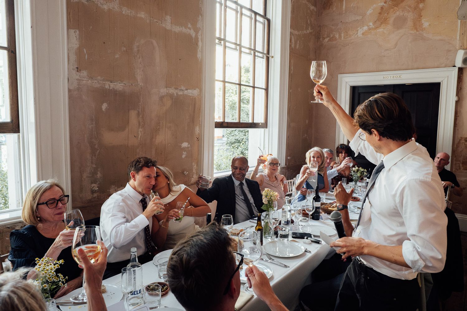 Guests seated around a long banquet table raise glasses as a man in a white shirt stands to give a toast in a rustic, sunlit dining room.