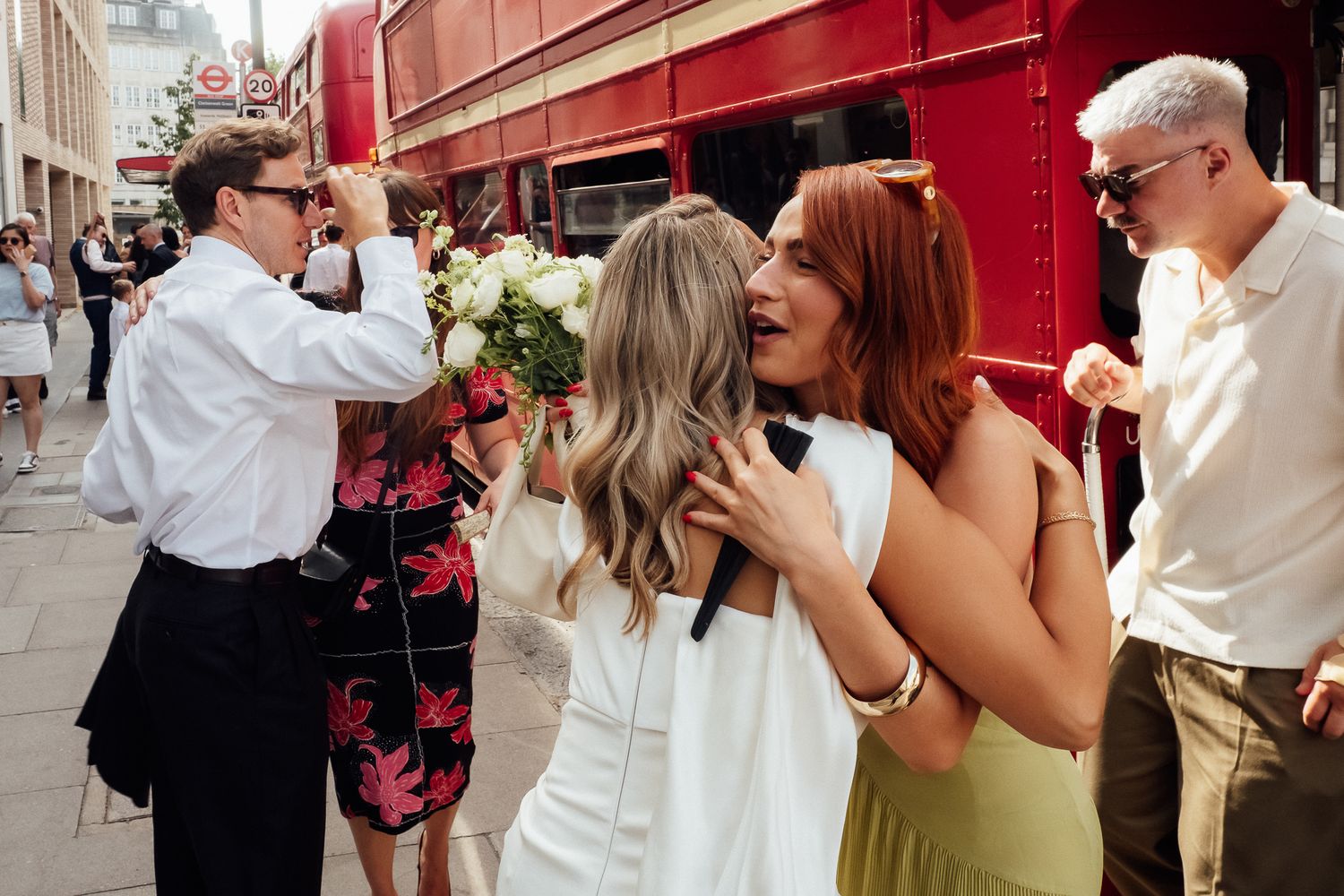 Two women in white and green dresses embrace and laugh near a red London double-decker bus while guests in sunglasses celebrate nearby.