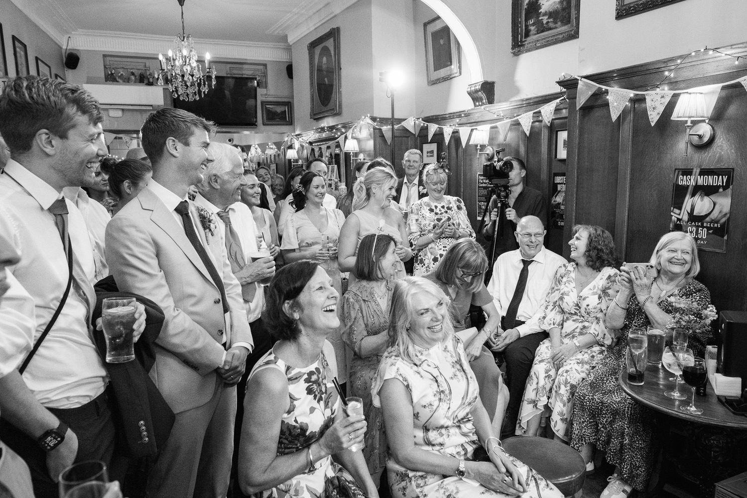 Smiling wedding guests in summer attire laugh together inside a warmly lit pub with a chandelier and bunting overhead.