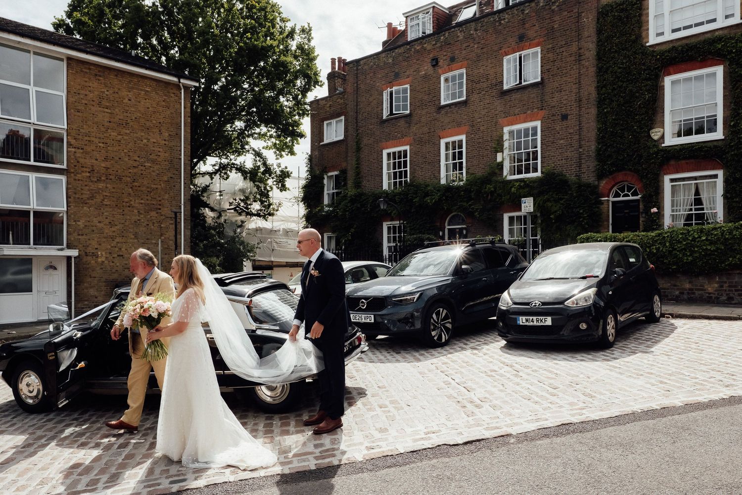 A bride in a white gown and veil walks with an older man holding a bouquet past parked cars outside a brick building.