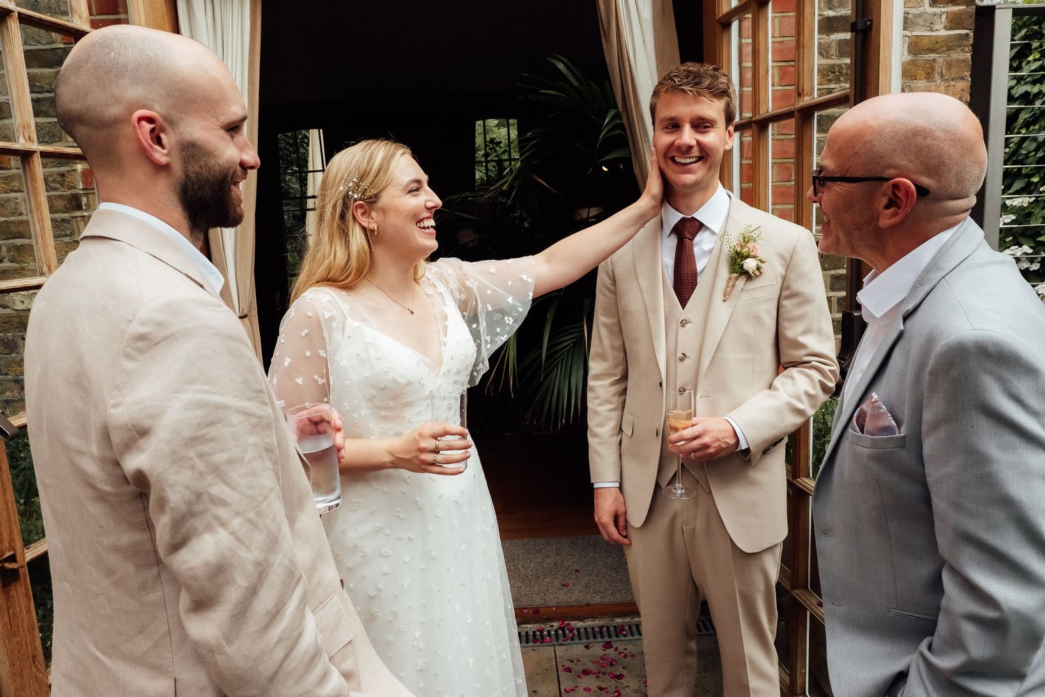 A smiling bride in a white lace wedding dress touches a groom's face while two men in beige suits stand beside them at a wedding.