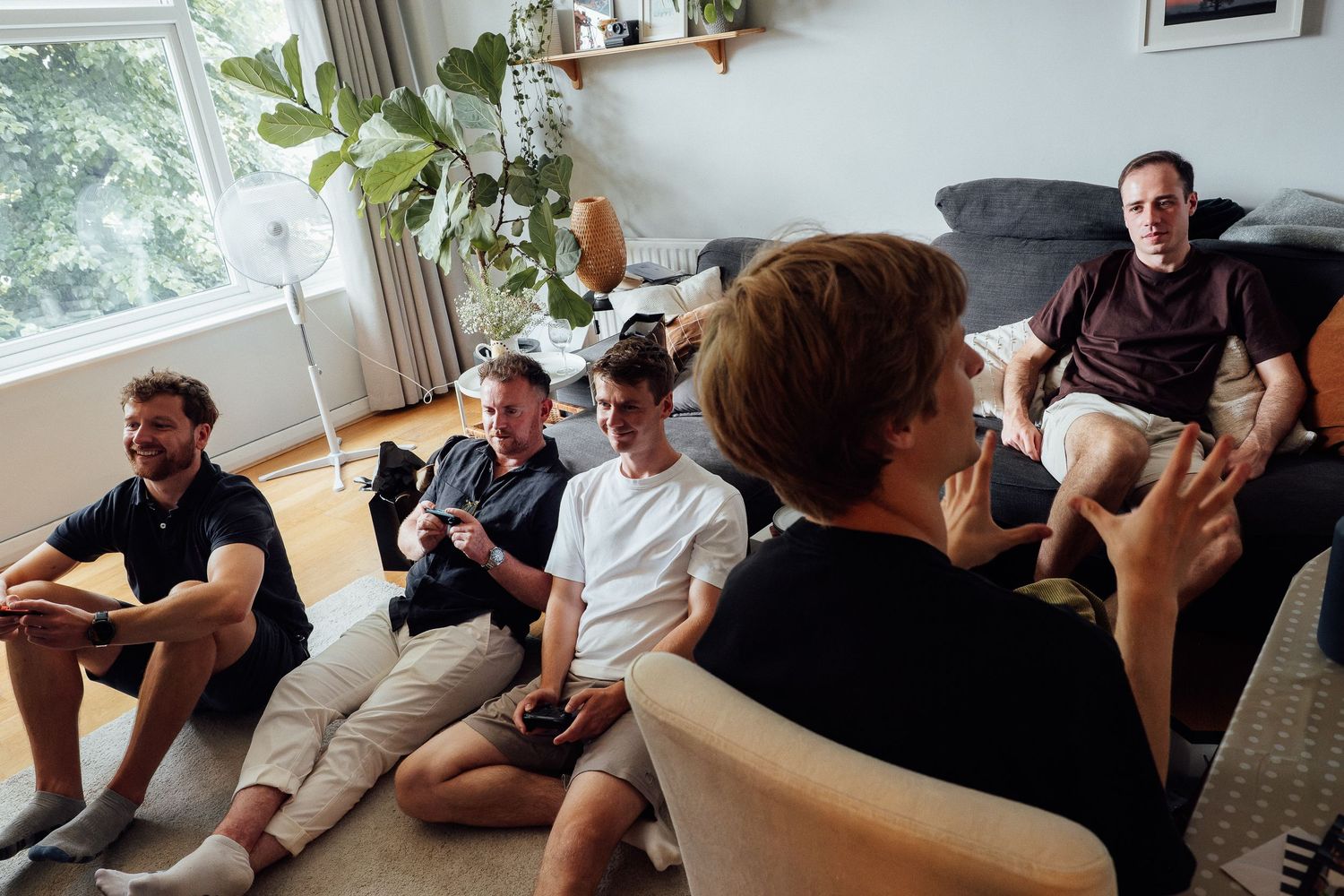 Five men relax in a bright living room, sitting on sofas and the floor while talking and holding video game controllers.