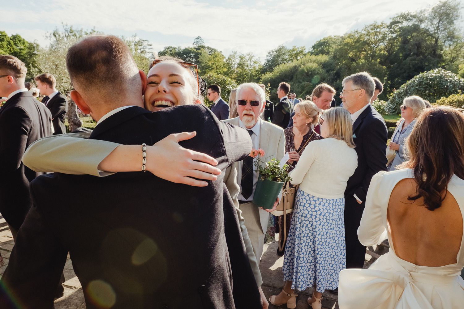 Wedding guests share joyful embraces during an outdoor reception celebration in a sunny garden setting.