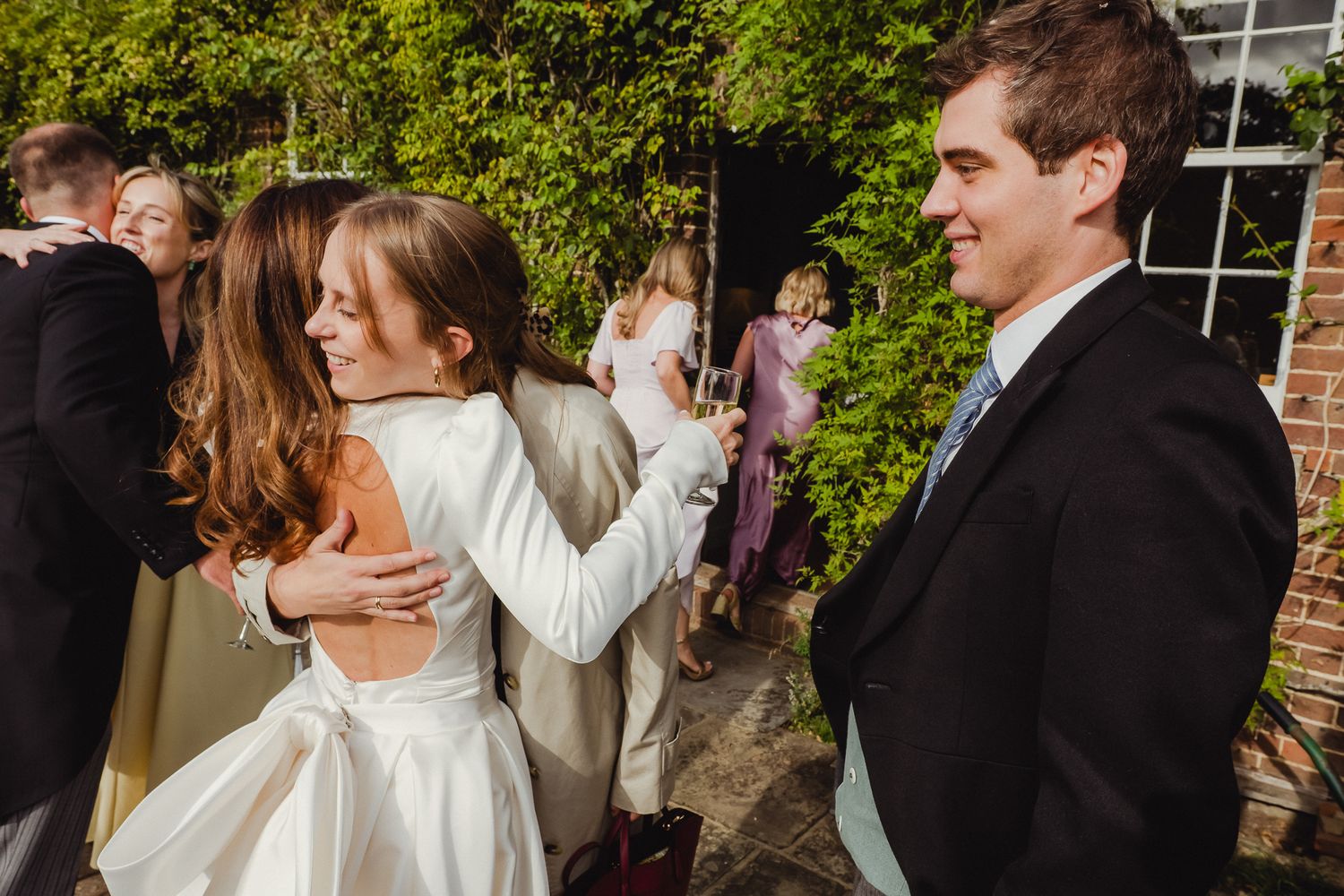 Wedding guests mingle and embrace during an outdoor reception by a vine-covered wall.