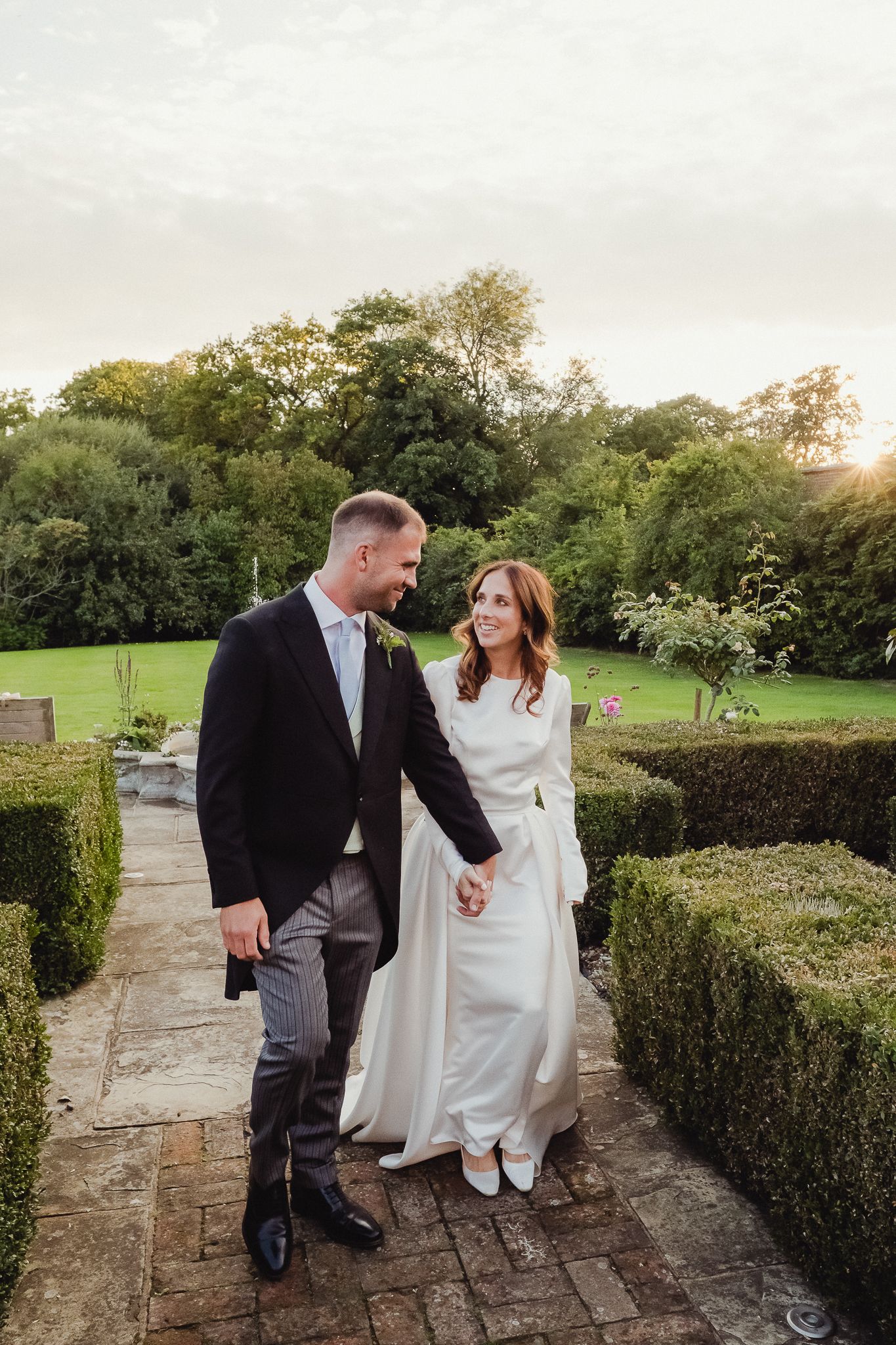 A couple in wedding attire walks along a stone path surrounded by manicured hedges and gardens at sunset.
