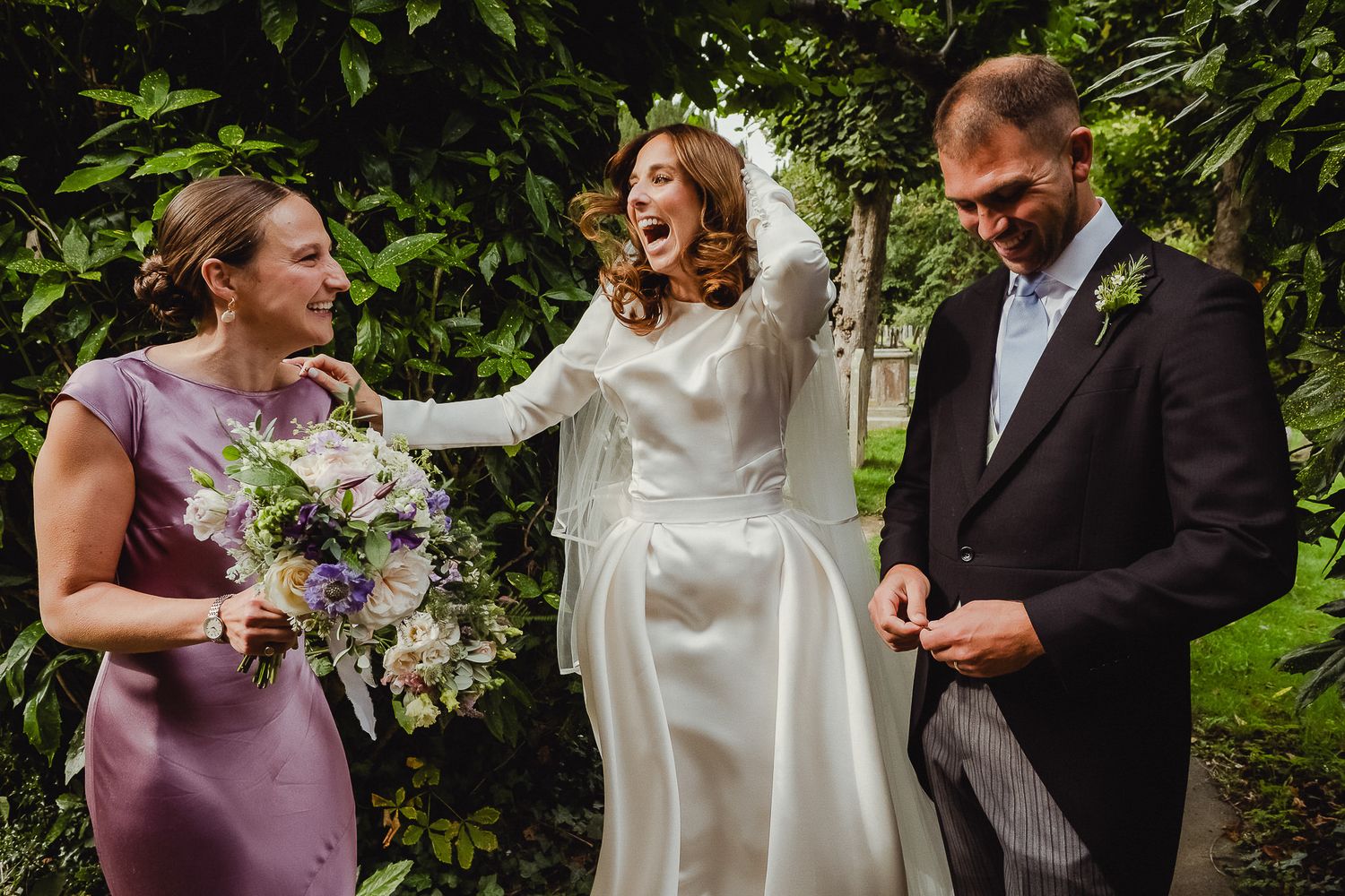 Wedding party laughs together during outdoor garden photo session with greenery in background.
