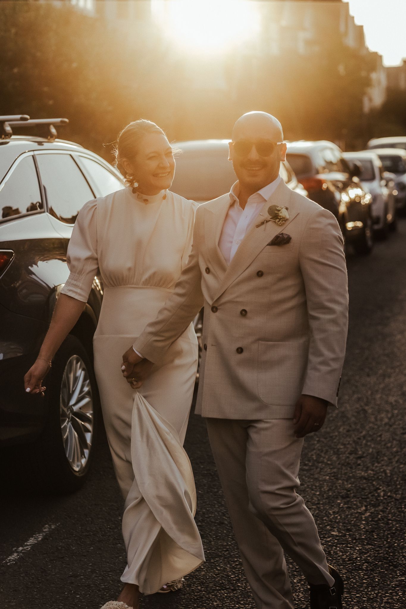 A couple in stylish wedding attire walk together at sunset, with parked cars visible in the background.