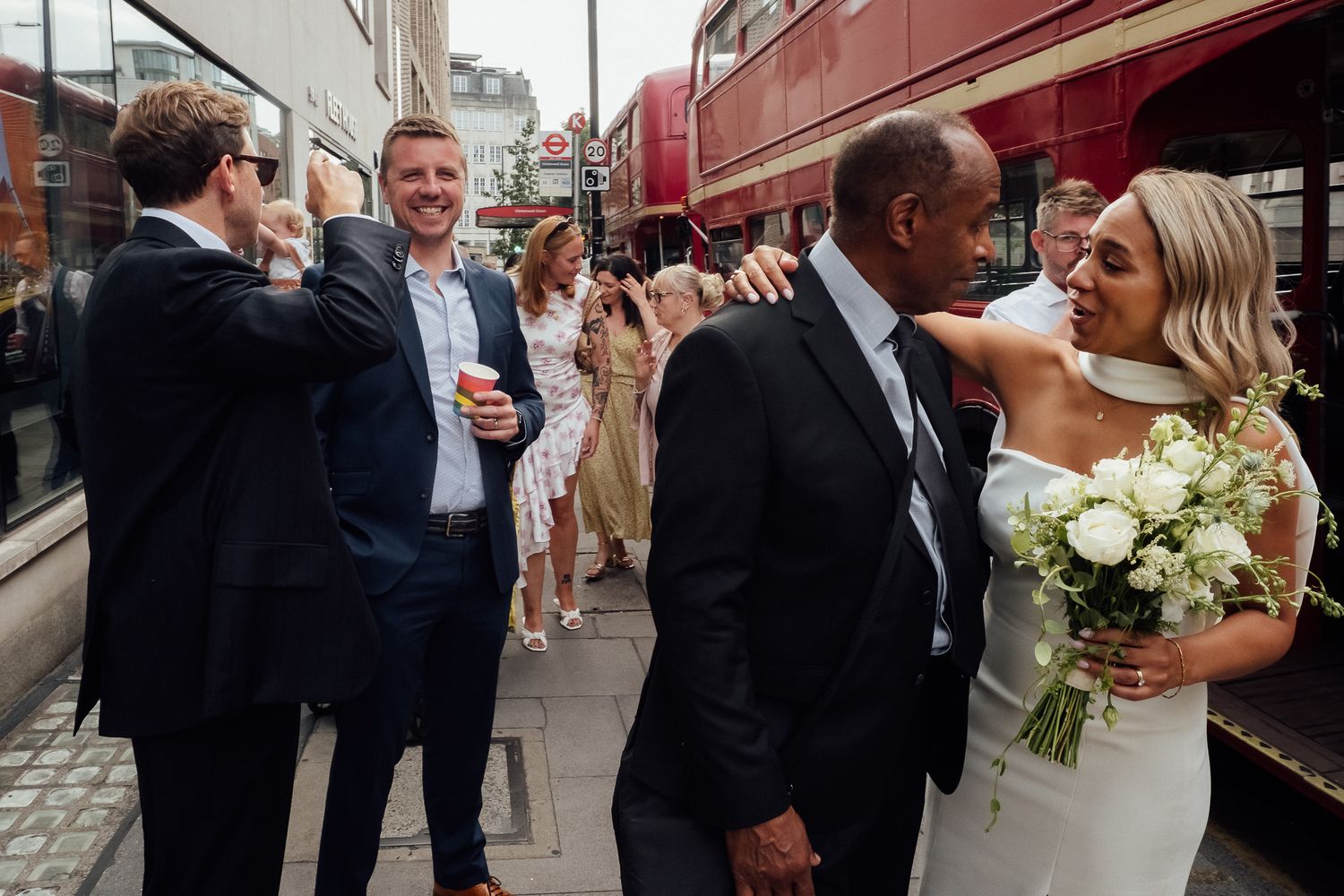 Bride in white dress holding a white bouquet embraces a man in a dark suit as wedding guests celebrate near a red London bus.