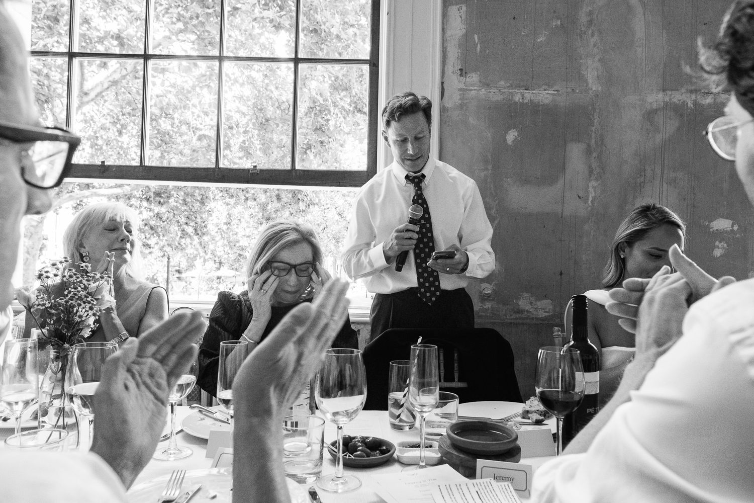 Man in a white shirt and tie stands giving a speech as seated guests applaud around a wedding dinner table with wine glasses.