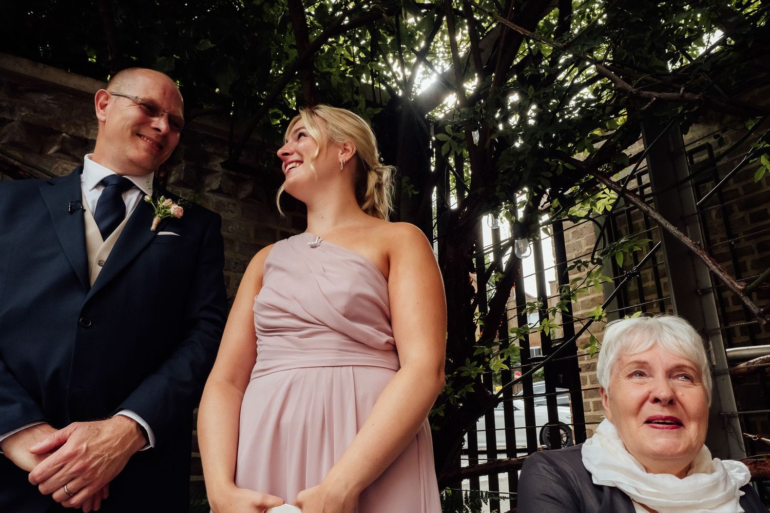 A smiling bridesmaid in a blush pink dress stands between a man in a navy suit and an elderly woman at an outdoor wedding ceremony.