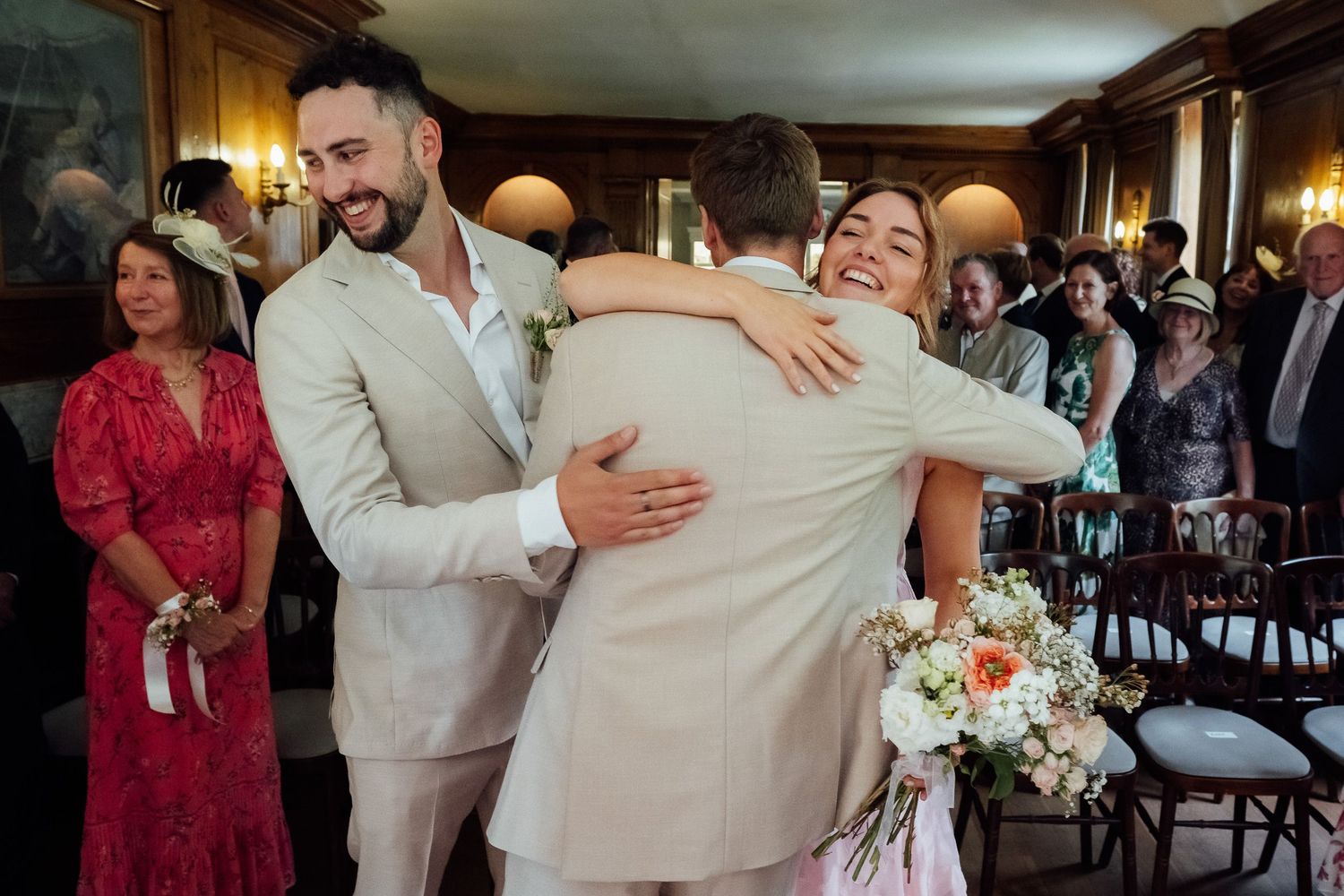 A smiling bride in a white gown embraces a man in a beige suit during a joyful wedding ceremony as guests look on.