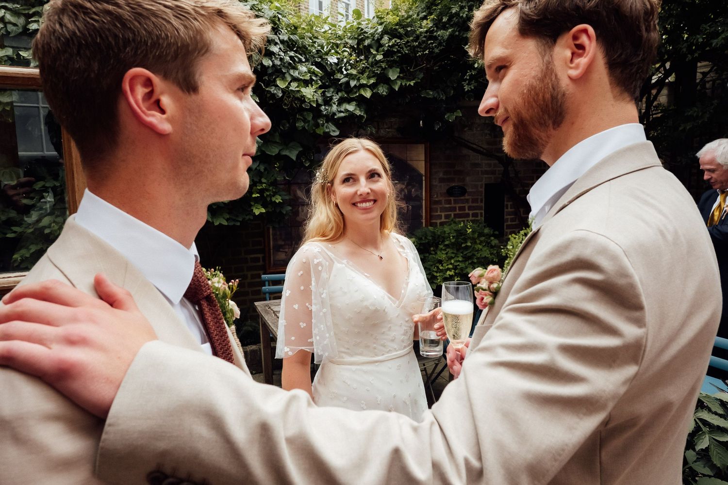 Two men in light suits greet each other warmly while a smiling bride in a white lace dress holds a drink in a leafy garden courtyard.