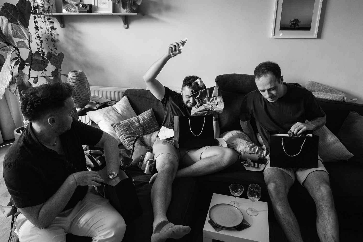 Three men relax on a dark sofa in a cozy living room, one reclining with a phone raised while another holds a tablet, in black and white.