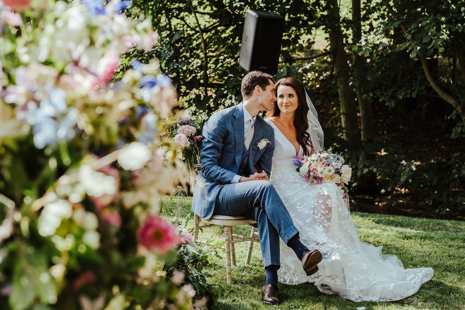 Newlyweds share an intimate moment while seated outdoors in a garden setting with floral decorations.