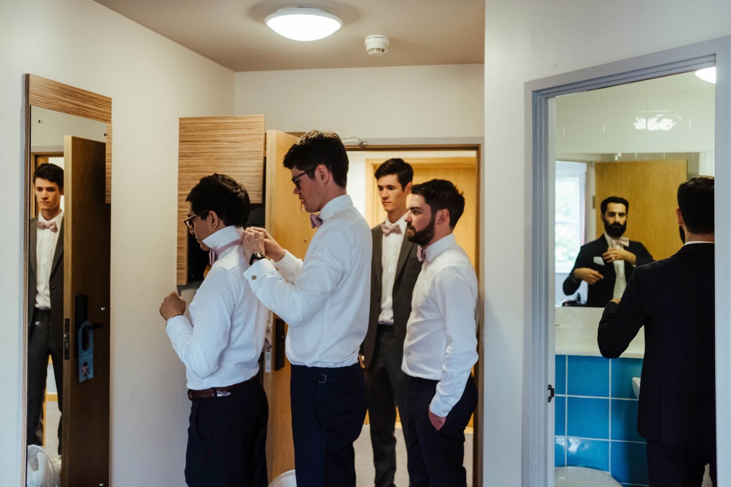 Groomsmen helping each other get ready in a dressing room with mirrors.