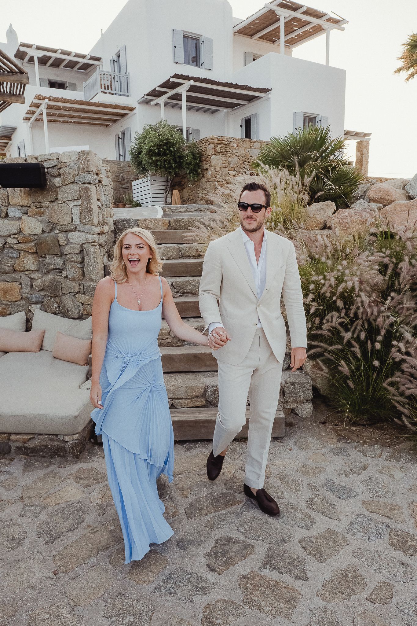 Couple walking down stone steps at a Mediterranean resort wearing blue dress and beige linen suit on a sunny day.
