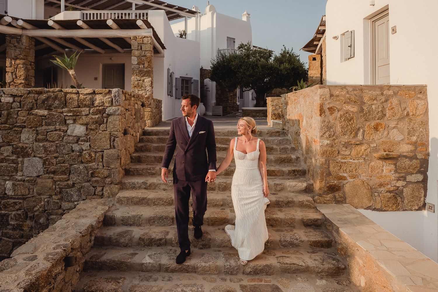 Two figures walk hand in hand between rustic stone walls at a Mediterranean destination during golden hour lighting.
