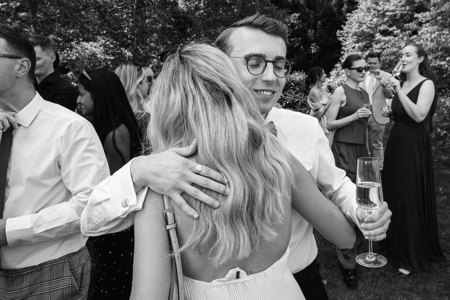 Black and white photo of a couple embracing at an outdoor wedding reception, man holding champagne glass.