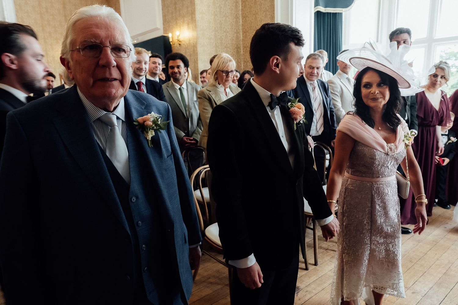 Elegant wedding ceremony with guests seated, featuring a groom in a suit and a woman in a lace dress and hat.