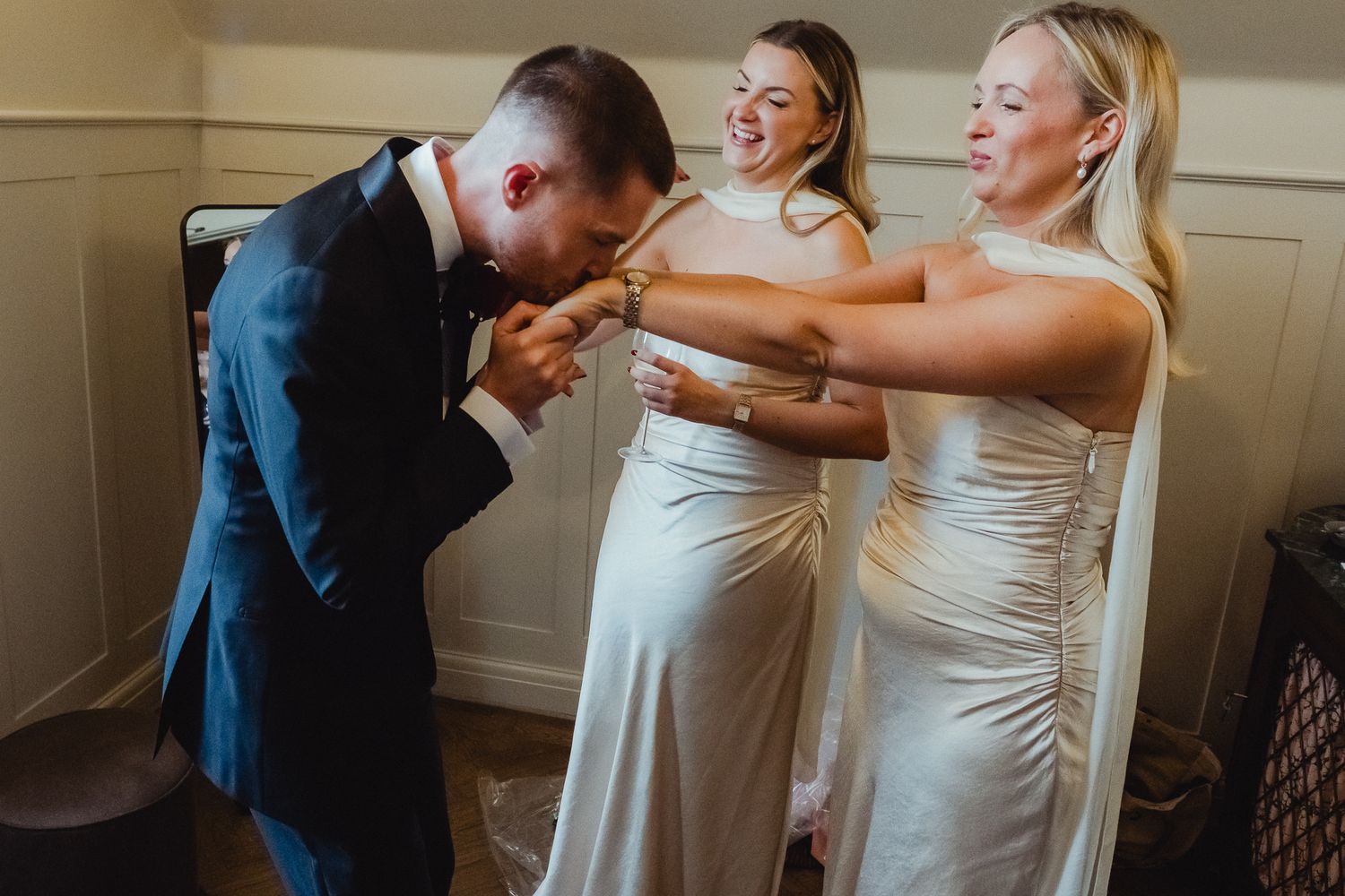 Two bridesmaids in white dresses share a joyful moment with someone in a navy suit during a wedding celebration.