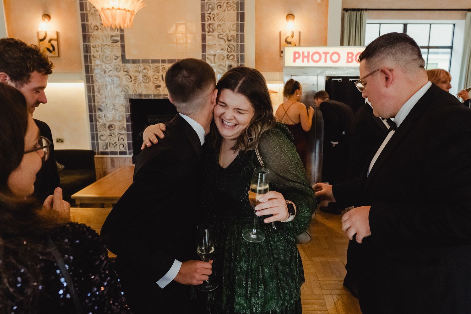Wedding reception guests socializing and laughing in front of exposed brick walls with decorative lighting.