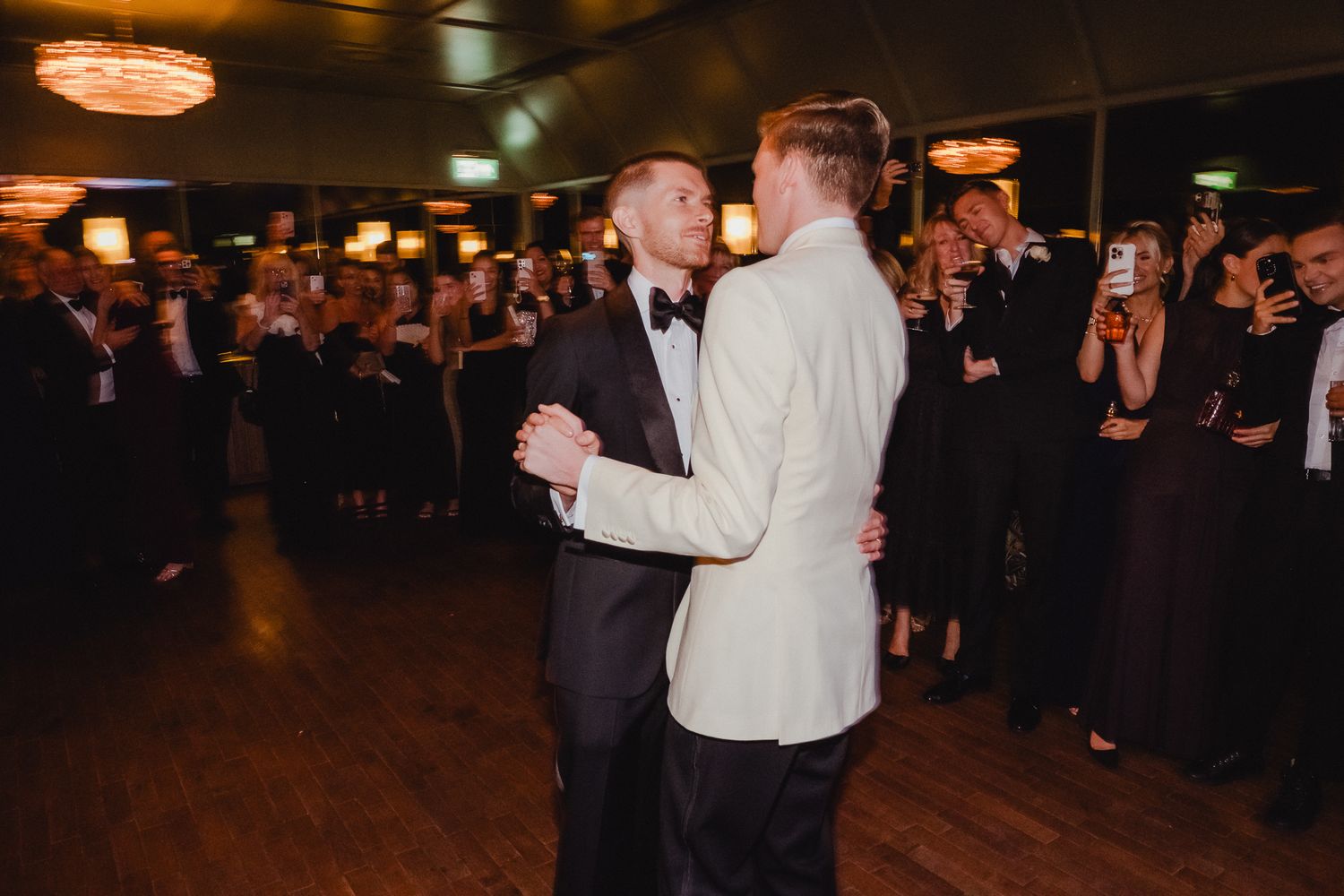 Two people share their first dance at a wedding reception while guests look on with phones capturing the moment.