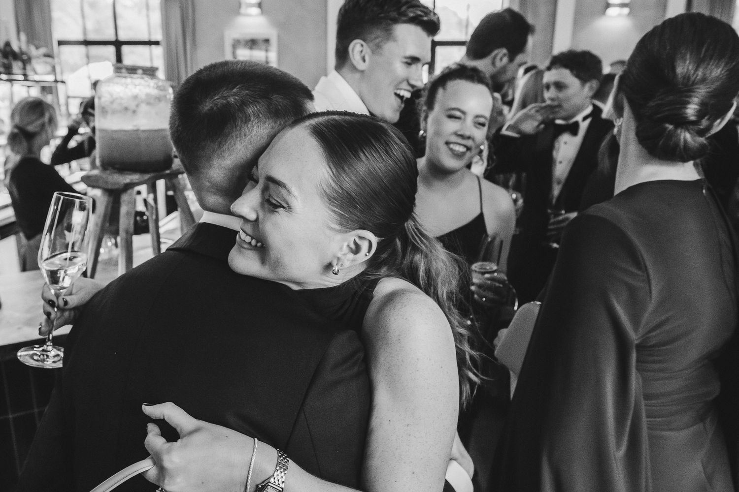 A crowded wedding reception shows guests mingling and hugging in a bright indoor space.