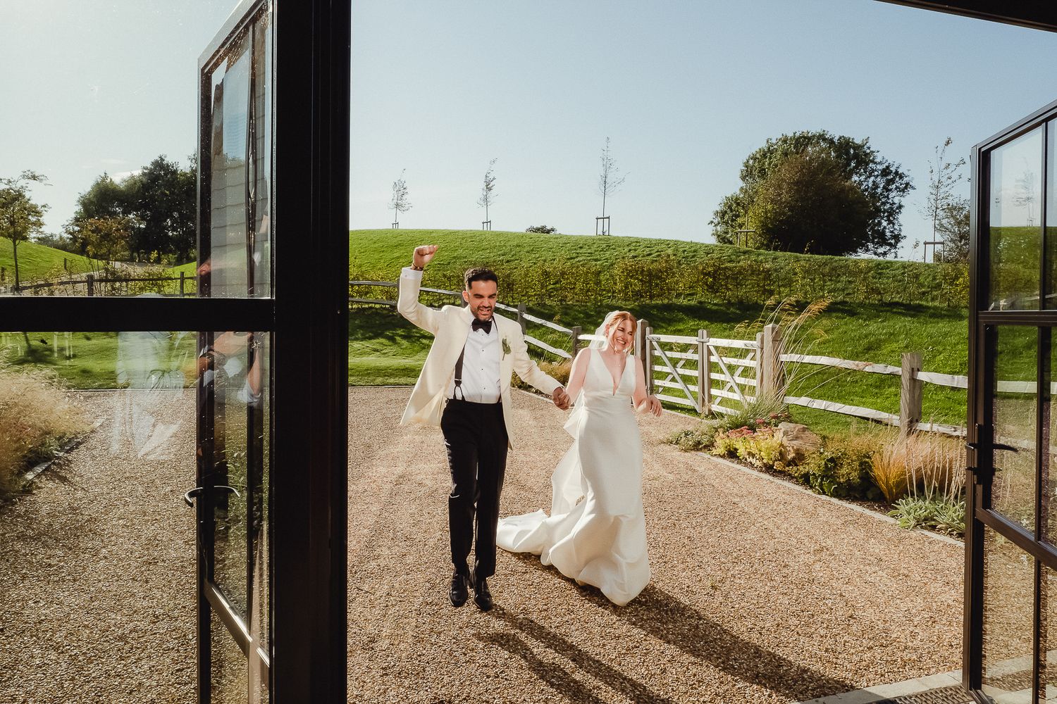 A couple in wedding attire walks along a gravel path surrounded by lush green countryside on a sunny day.