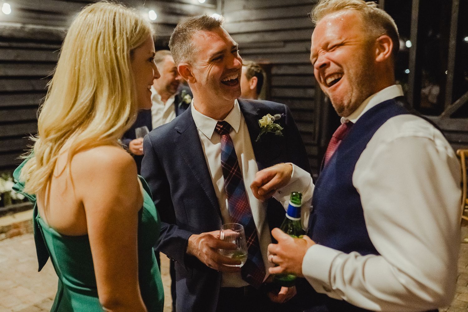 Three people share a candid laugh during a wedding reception, with one wearing a green dress and others in formal attire.