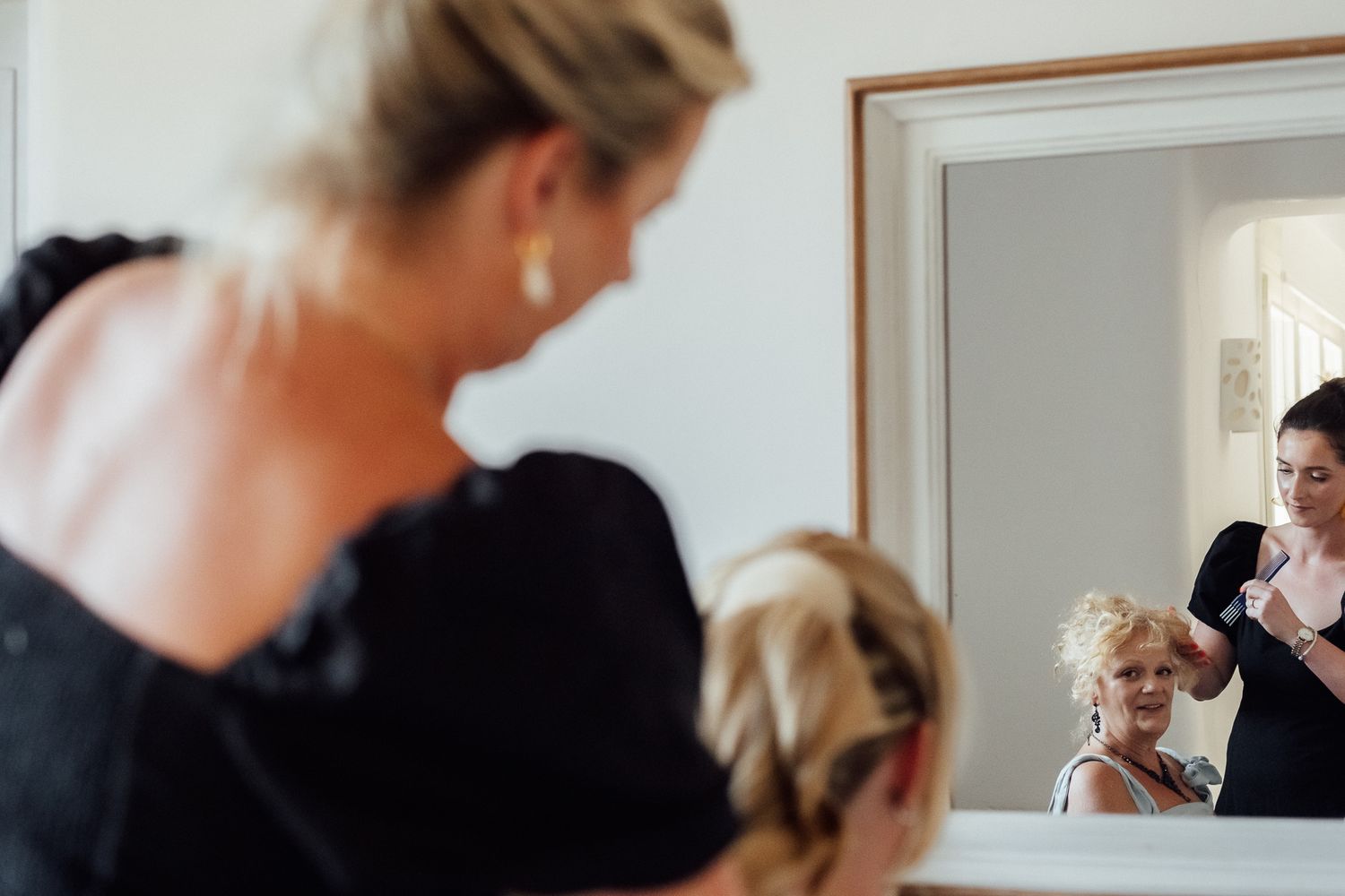 Woman styling blonde hair reflected in mirror, wedding preparation scene with elegant black dress.