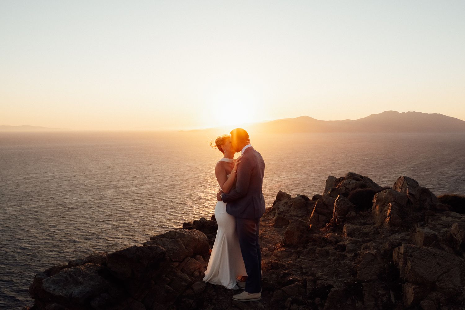Couple embracing at golden sunset on rocky cliff overlooking ocean, wedding attire, romantic elopement photography.
