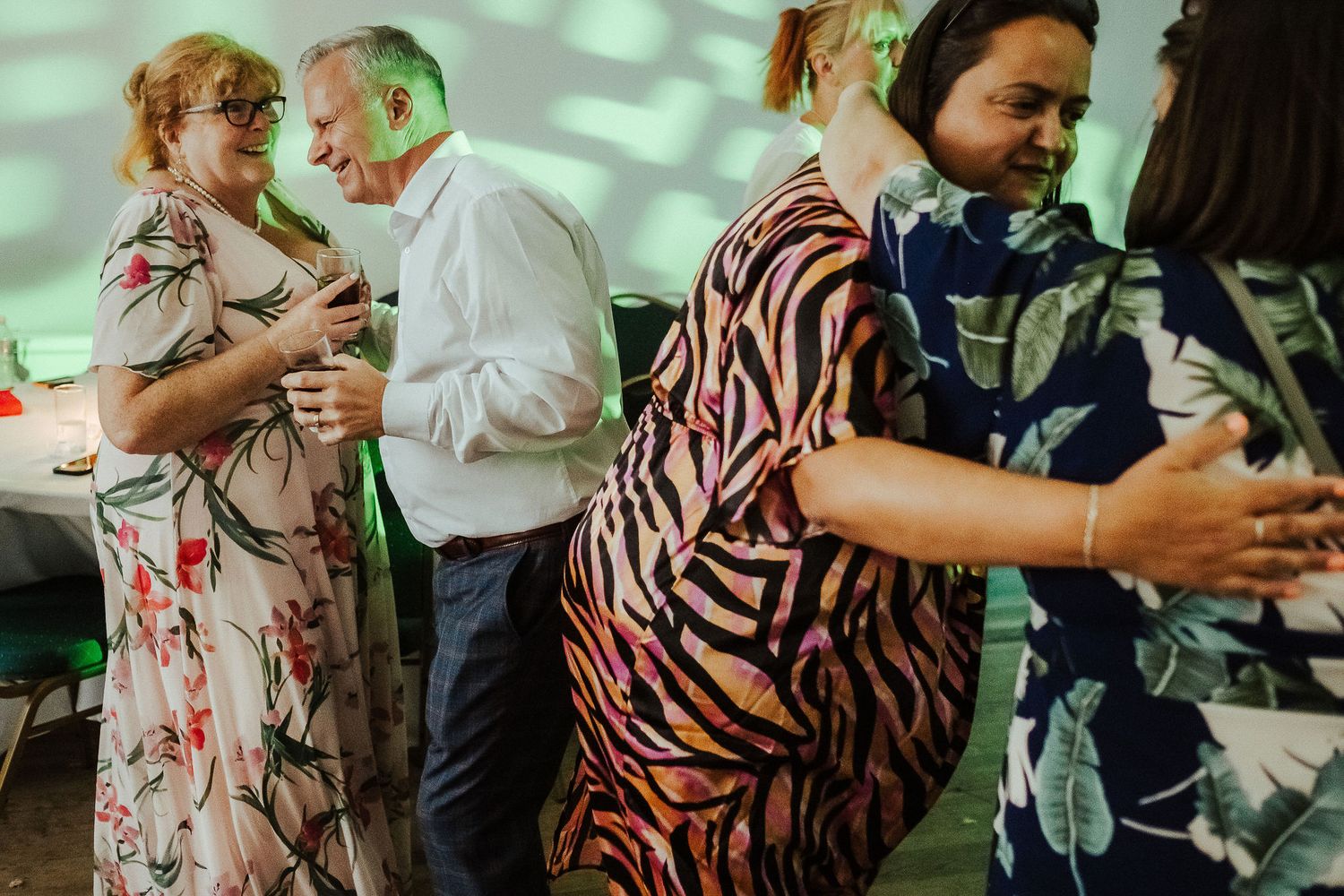 Guests in floral and zebra-print outfits dance and chat at a lively indoor party with soft green lighting.