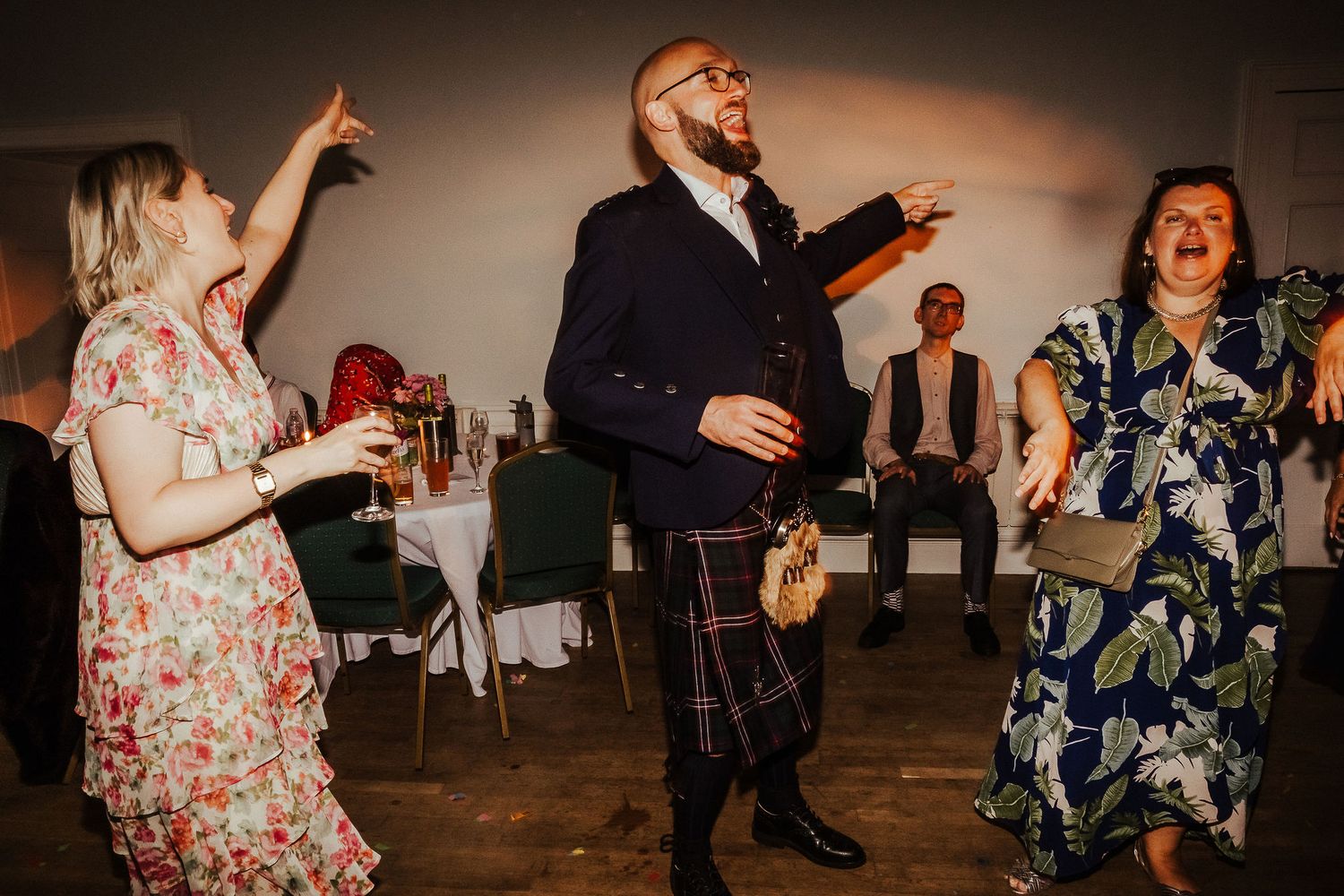 Wedding guests dance joyfully in a reception hall, including a bearded man in a navy jacket and tartan kilt and women in floral dresses.