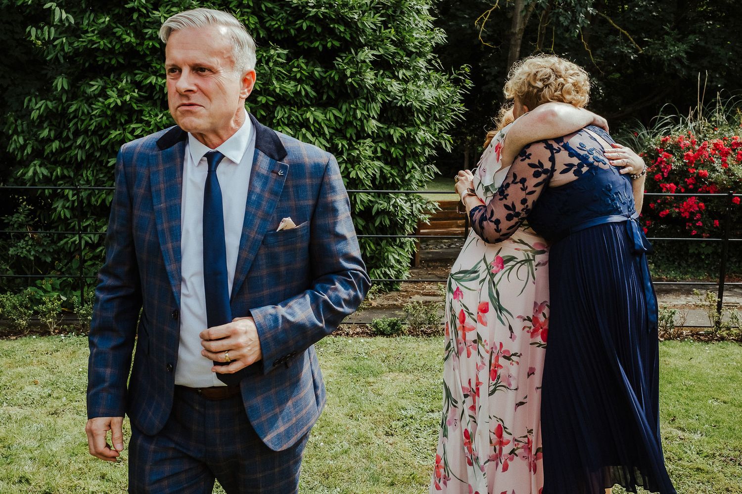 Older man in a blue plaid blazer stands in a garden while two women in floral and navy dresses embrace behind him.
