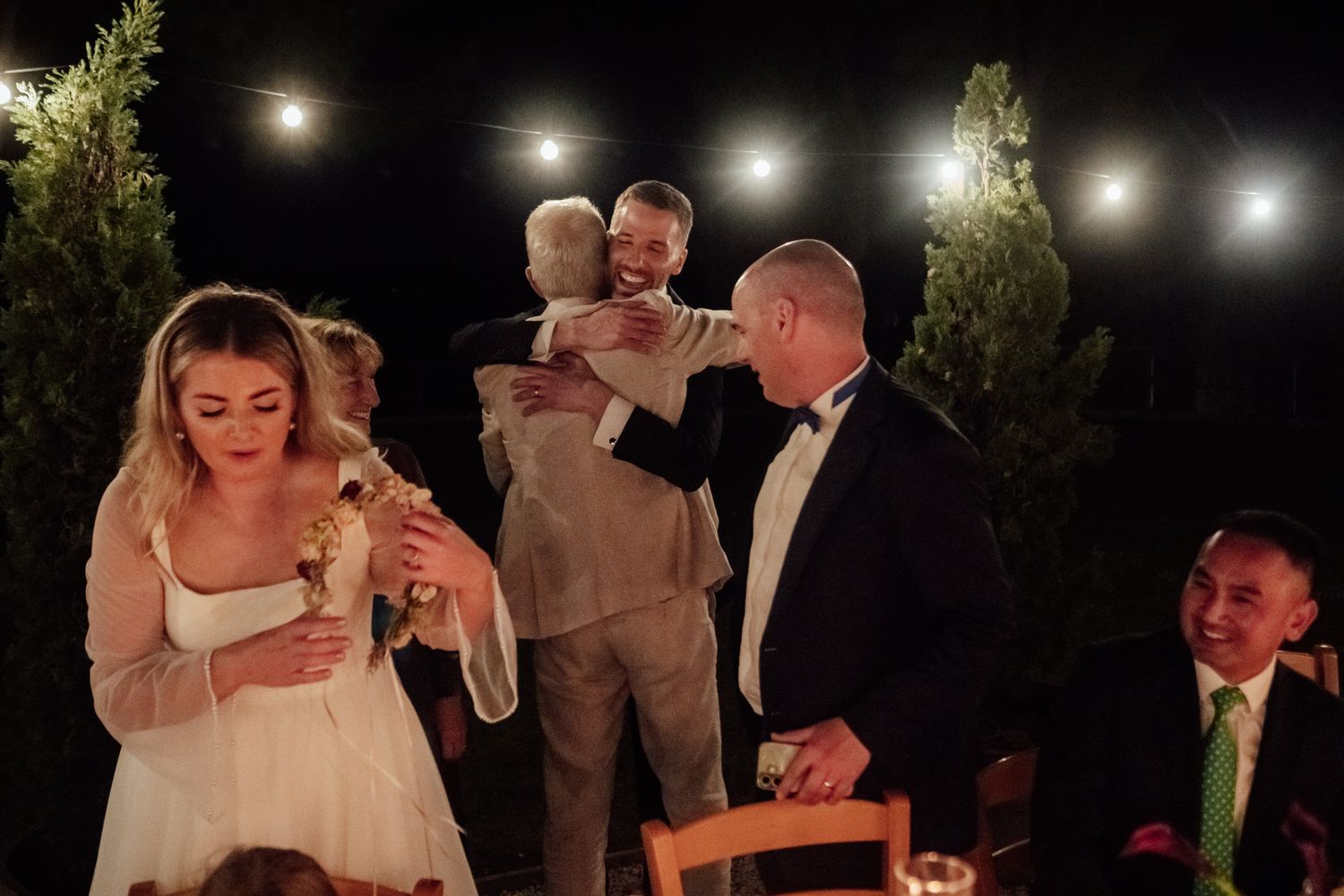Two men embrace at an evening wedding reception under string lights while a bride in a white dress and a smiling guest look on.