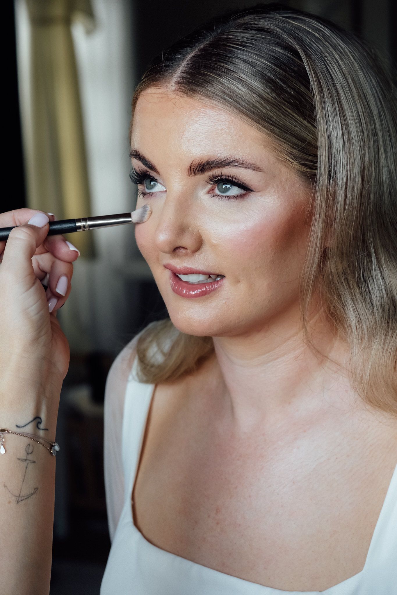 Makeup artist applies concealer with a brush to a woman with long blonde hair and glowing skin, wearing a white square-neck top.