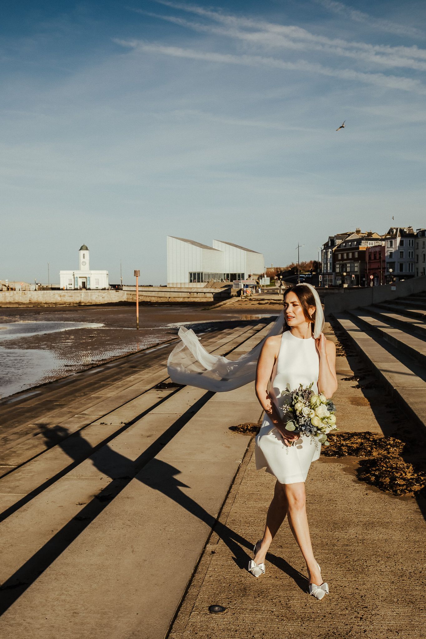 A coastal wedding photo series showing a person in white holding a bouquet against a lighthouse and beach backdrop at sunset.
