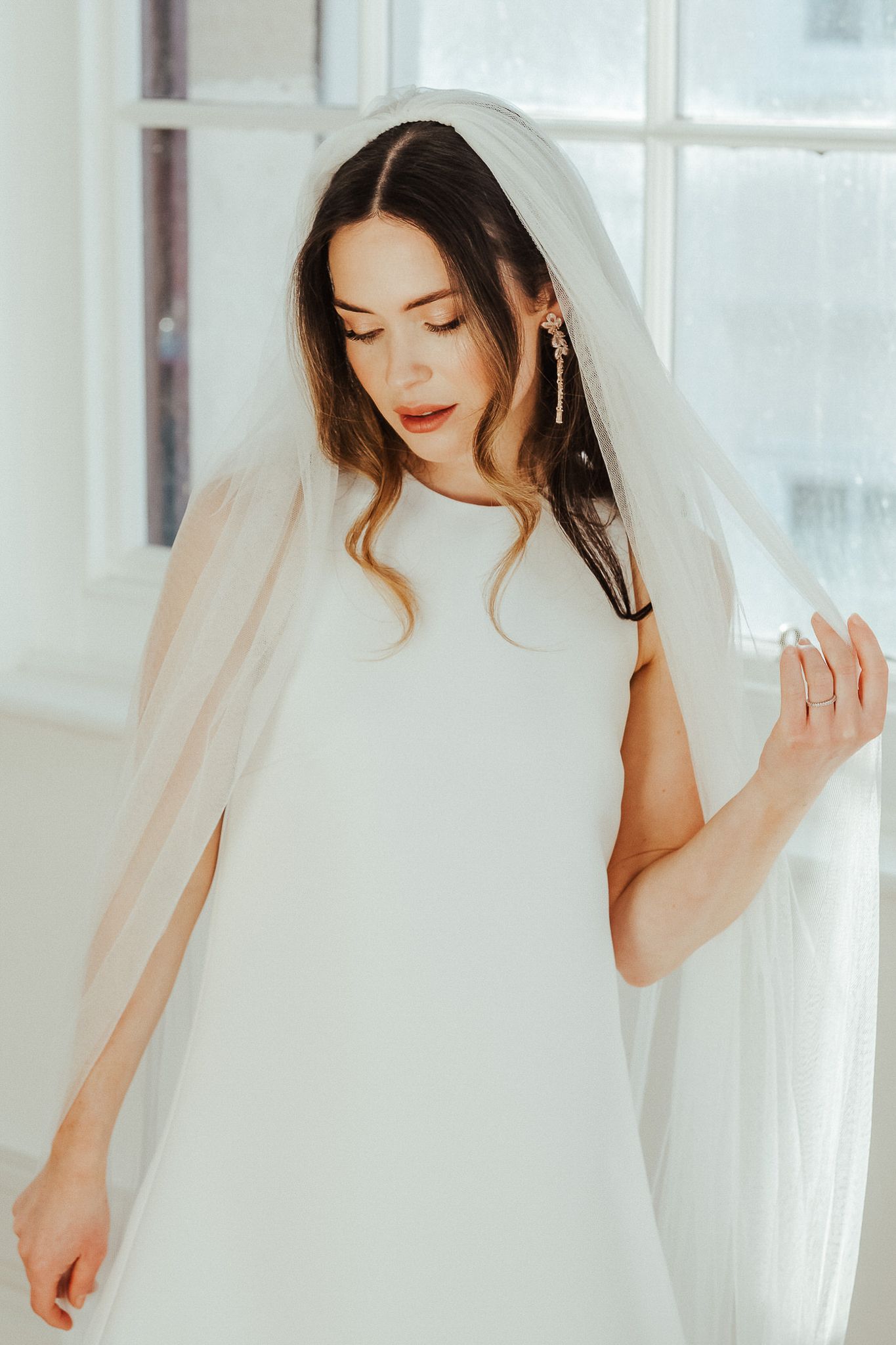 A bride in a flowing white veil poses gracefully against bright windows in a romantic bridal portrait series.