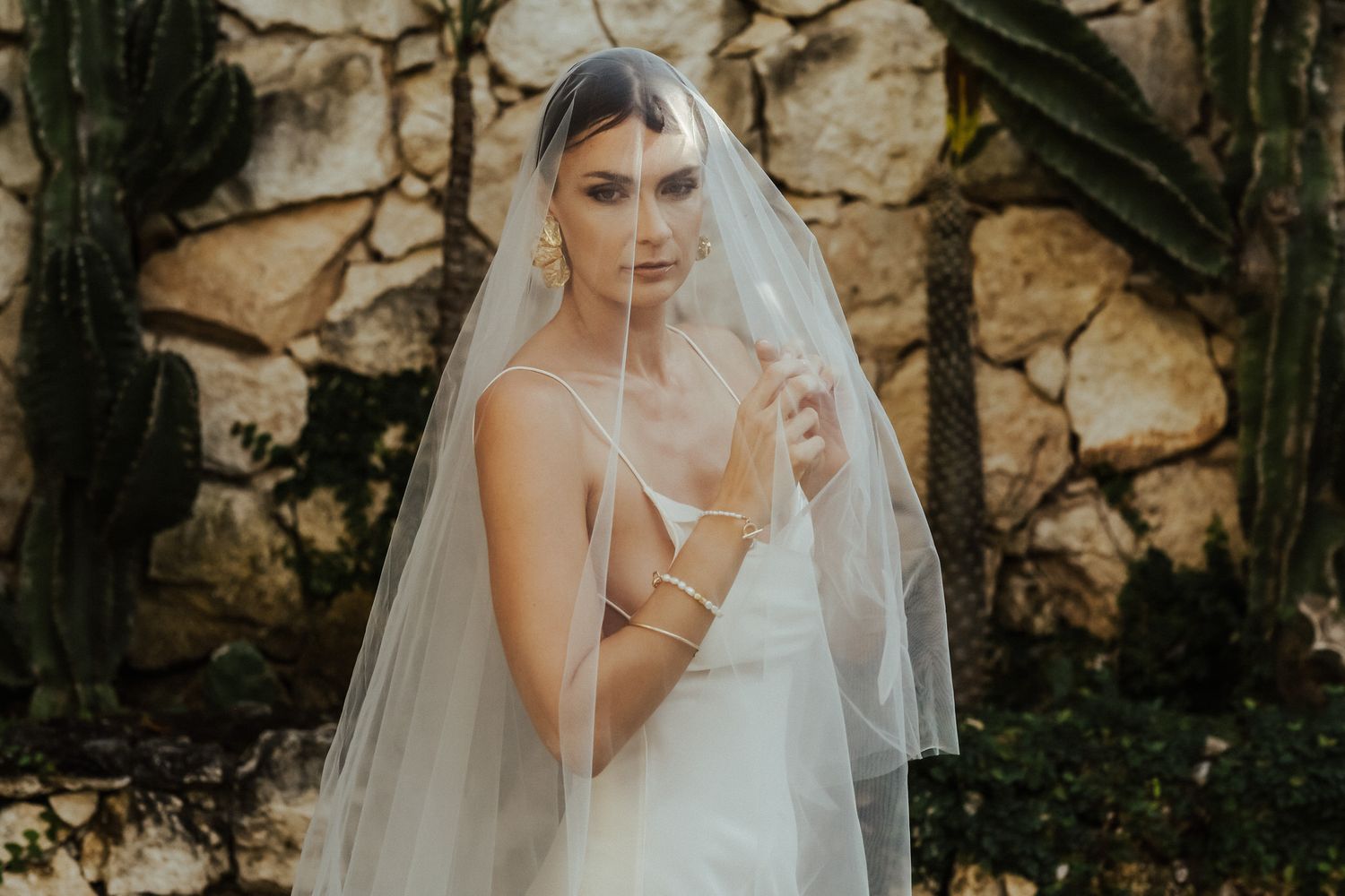 A bride in a flowing white gown and veil poses against a rustic stone wall backdrop in a romantic garden setting.