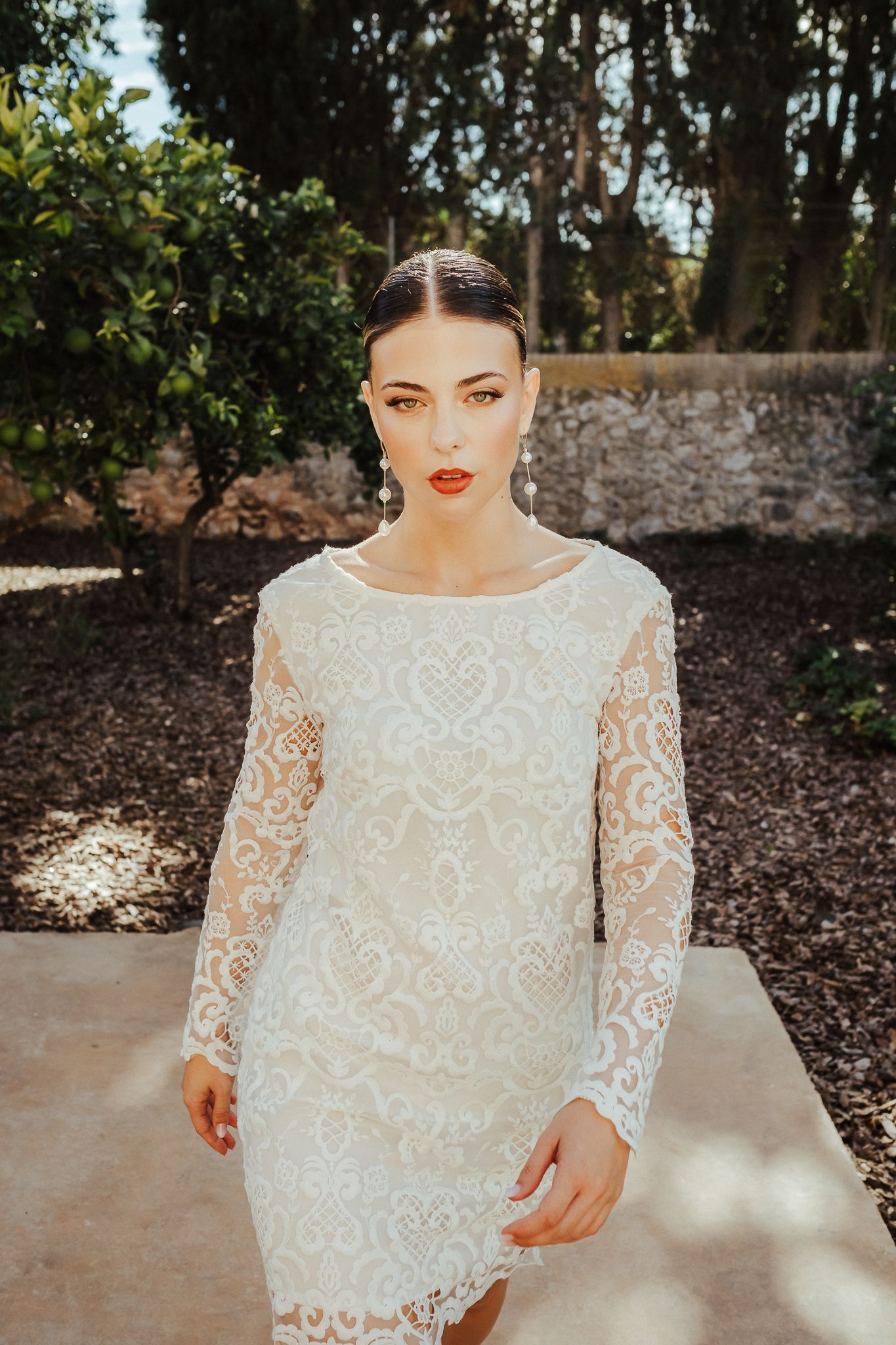 A bride in a white lace wedding dress with long sleeves and bold red lipstick poses in an outdoor garden setting.