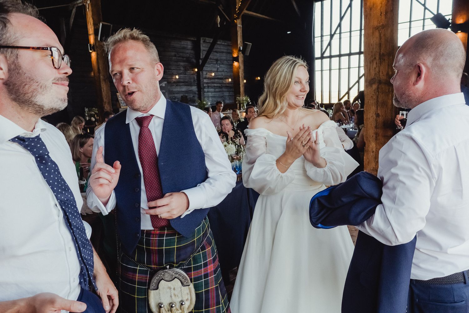 Wedding guests in traditional Scottish attire chat and laugh during a lively reception celebration.